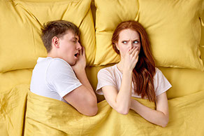 Ein Paar liegt im Bett. Die Frau hebt sich die Nase zu und schaut leicht angewidert auf den offenen Mund ihres schlafenden Partners.